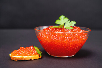 National Russian holiday delicacy - red salmon caviar in glass bowl and on piece of bread decorated with fresh parsley, black background, front view.