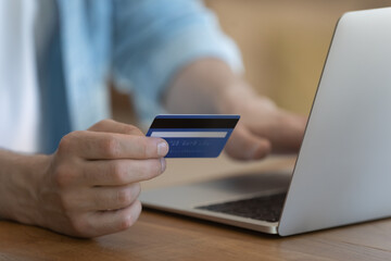 Close up young man holding banking card in hands, entering payment information or cvv code enjoying safe online shopping on computer, easy technology usage, electronic wallet, virtual money concept.