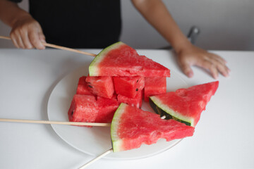 a child's hand holds a slice of ripe watermelon on a wooden stick, next to it is a plate with square slices of watermelon. Image with selective focus