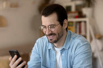Obraz na płótnie Canvas Head shot emotional positive young handsome guy in glasses holding smartphone, reading message with good news. Pleased millennial bearded man enjoying using mobile applications alone at home.