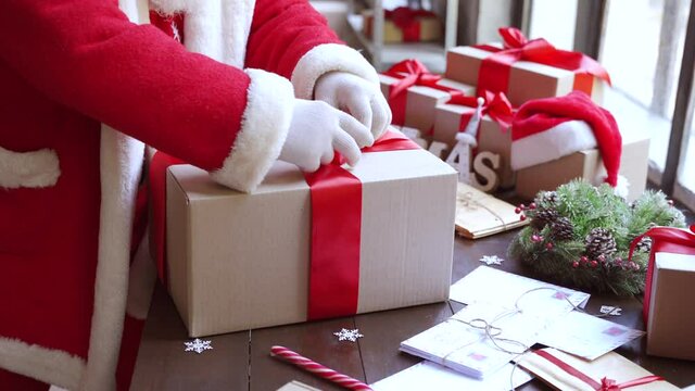 Closeup view of Santa Claus, Saint Nicholas packing present box wrapping gift with red ribbon preparing post shipping delivery parcel on table in workshop. Merry Christmas shipping delivery concept.