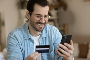 Head shot young smiling man in eyewear holding banking card in hand, entering security payment information in mobile application, purchasing goods or services, people and online shopping concept.