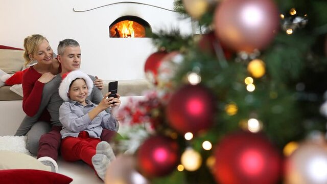Merry christmas, happy family at home, parents with son take a selfie with mobile phone near the illuminanted and decorated tree, joyful and smiling sitting in living room and comforted by fireplace 