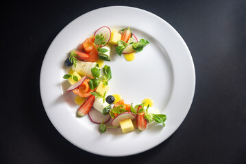 Red fish, vegetables and fruit salad on a white plate. Fish fillet rolls, radish, zucchini rolls, microgreens, strawberry wedges, mango cubes, juice drops. Top view, on a dark black background.