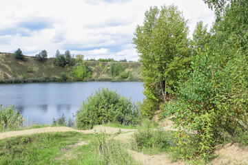 Summer landscape. The shore of a small lake with paths, trees, bushes. Cloudy sky