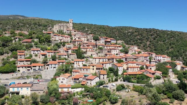 Scenic aerial view of compact village of Eus on green slopes of hill with ancient church of St. Vincent and stone castle ruins on hilltop, France