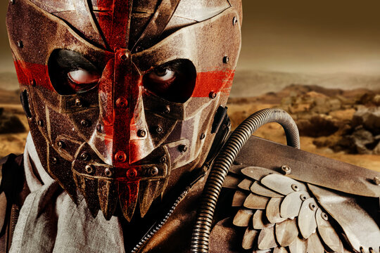 Photo of a post apocalyptic raider warrior in leather jacket with metal armor and steel mask with red cross painting standing in desert wasteland.
