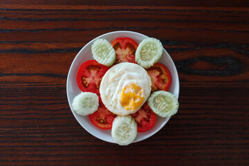 Fried egg served with slice fresh tomato and cucumber on white plate and wooden table