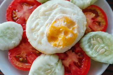 Fried egg served with slice fresh tomato and cucumber on white plate and wooden table