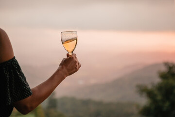 Woman's hand holding glass of white wine across beautiful mountains landscape and sunset