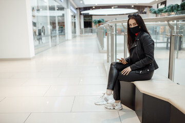 Girl with medical black mask and mobile phone in a shopping center. Coronavirus pandemic. A woman with a mask is standing in a shopping center. A girl in a protective mask is shopping at the mall