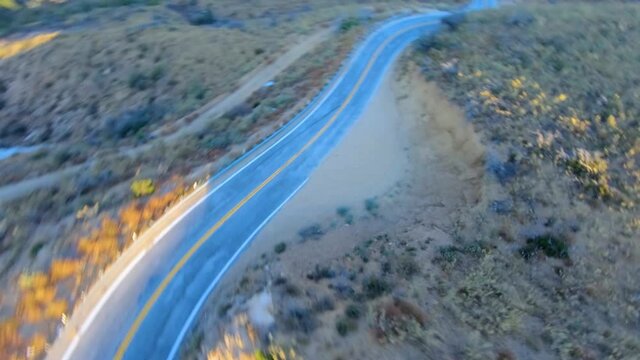 Aerial FPV, drone shot tracking a blue car driving on a curvy mountain road, during sunset, in Los Angeles, California, USA - First person view