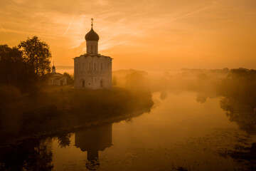 Aerial shot of a Russian Orthodox church above a misty field lit by the rising sun. A pond near the church, a river in the background. No people.