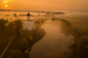 Fototapeta premium Aerial shot of a Russian Orthodox church above a misty field lit by the rising sun. A pond near the church, a river in the background. No people.