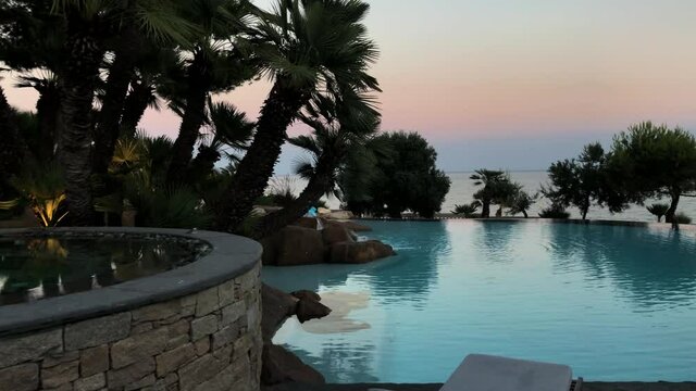 View of an idyllic garden with infinity pool, palms and trees next to the sea. View of a jacuzzi and a swimming pool in a sunset. Panning right over a swimming pool.