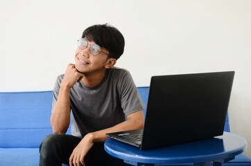 young Asian man sitting on the sofa in front of the laptop with an expression of finding ideas