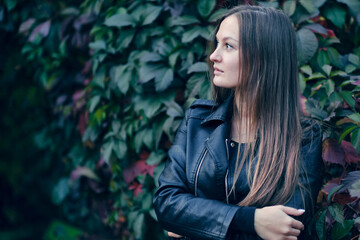 Fototapeta na wymiar young beautiful woman in a stylish black leather jacket looks to the side, against the background of wild grapes. Autumn portrait in dark tones.