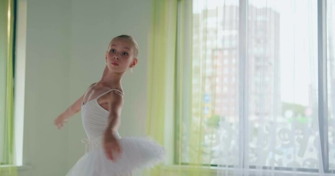 Concentrated little girl in white suit dances doing classical ballet spins at window with curtain in studio closeup slow motion