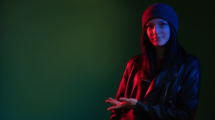 Night portrait. Neon light. Street style woman. Information banner. Confident lady in hat leather biker jacket in red blue glow showing copy space isolated on dark green advertising background.