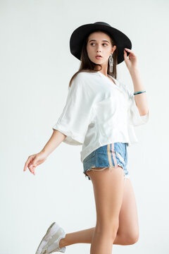 Summer look. Hipster style. Beautiful young woman in white blouse jeans shorts straw black hat combining sportive casual clothes isolated on neutral. Fashion posing. Trend outfit