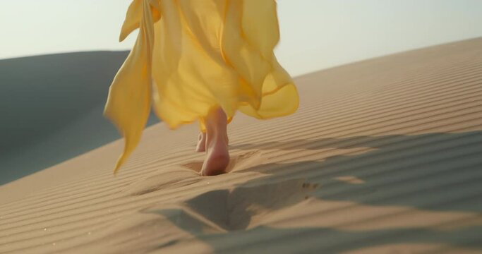 Fashionable shot: close up view of slim woman legs walking by the rippled sand dune surface. Female model in bright yellow flattering dress walking toward horizon. 4K stylish clothes is waving on wind