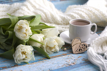 on a blue wooden background: a bouquet of white tulips, a cup of coffee, a carved wooden heart, a white knitted scarf. Greeting card for Valentine's Day, on Mother's Day, on March 8.