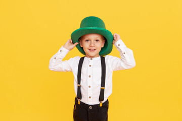 Smiling little child boy in green leprechaun hat on yellow background. St. Patrick Day celebration. Funny face