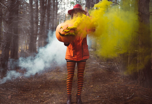 Girl in the orange jacket and striped stockings with heels in the autumn forest holds a large pumpkin for Halloween with yellow smoke.