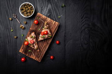 Appetizer bruschetta with tuna, capers and tomatoes on wholemeal bread. Italian cuisine, Delicious breakfast or snack on a dark background top view