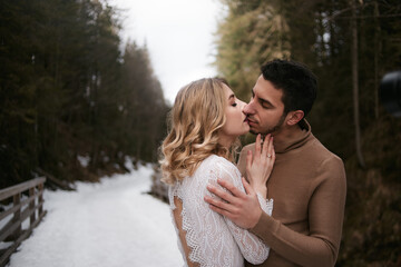 newlyweds bride and groom kissing in snow park during winter. woman wears minimalist white wedding dress. man wears casual clothing