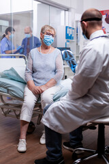 Medical specialist in hospital room examining x-ray of old woman and giving a diagnosis while wearing protection mask against coronavirus as safety precaution. Check up for infections, disease .