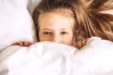 Child hiding under duvet in bed. Morning routine. Indulge in the bedroom in the morning. Girl in bed look out from under white covers. Selective focus