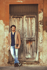 Obraz na płótnie Canvas Handsome young man is leaning against the wall near the door of an old house entrance. A ruined wooden door.