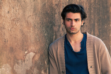 Engaging handsome man and old wall. Beautiful hair style. A charming Italian guy leaning against the wall. Stylish hair, beauty and attractive look. Serene and male gaze. Much copy space to the left.