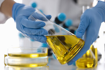 Hands in rubber gloves hold flask with yellow liquid in chemical laboratory closeup. Quality...