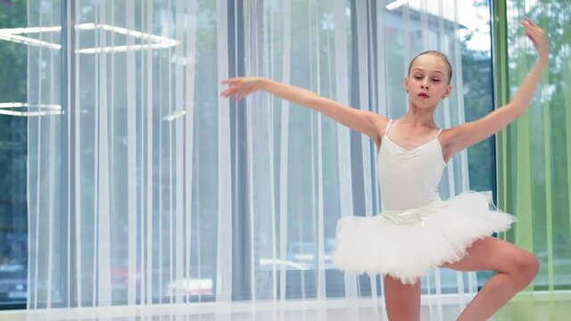 Talented little ballerina in white tutu dances classical ballet near window with curtains in spacious studio slow motion closeup