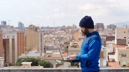 Portrait of a hipster man with hat working with a laptop on a rooftop in a city	