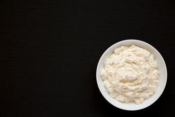 Tasty Ricotta Cheese in a white bowl on a black background, top view. Overhead, from above, flat lay. Copy space.