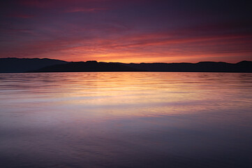夕暮れの湖。穏やかな波の水面に映る空の色。遠くの山のシルエット。