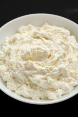 Tasty Ricotta Cheese in a white bowl on a black surface, side view. Close-up.