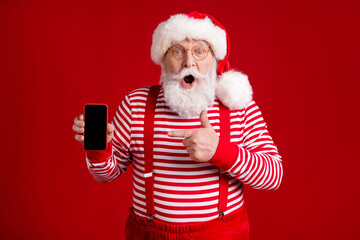 Photo of retired old man grey beard shock hold telephone direct finger screen empty space wear santa x-mas costume suspenders spectacles striped shirt cap isolated red color background