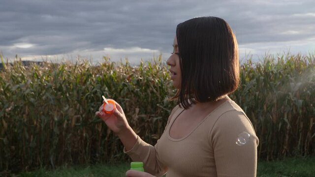 Slowmotion footage of brunette girl blowing soap bubbles beside scenic field and cloudscape at sky.