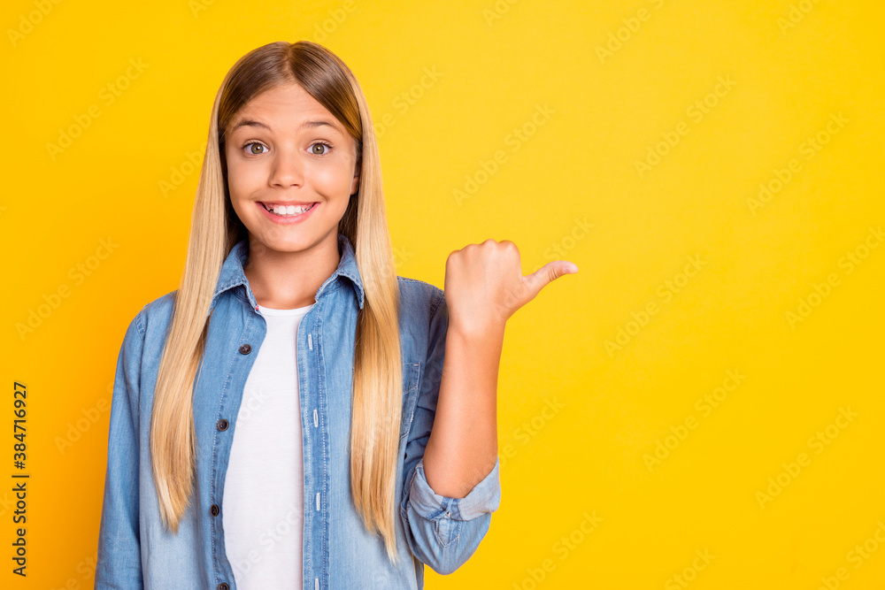 Poster Photo portrait of blonde schoolgirl showing with thumb on empty space smiling isolated on bright yellow color background