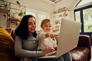 Little toddler girl sitting on mother lap using laptop on couch and pointing on screen showing something