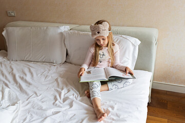 Cute little caucasian girl in pyjama reading a book. Kid sitting on a bed in the room. Stay at home during coronavirus covid-19 lockdown.