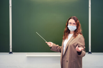 Teacher in face mask shows with pointer on school blackboard, copy space. Concept of problems at school during the coronavirus epidemic