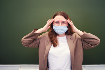 A teacher in a medical mask stands with a headache at the blackboard. Education during the...