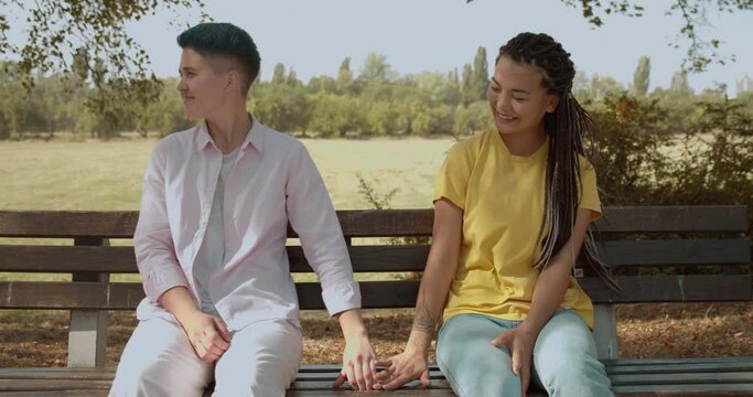Happy lesbian couple holding hands, sitting on the bench in park, first date