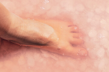 Fototapeta premium Woman leg foot close-up in pink salt flakes top view under pink water surface. Healthy spa procedures
