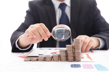 A male investor in a black formal suit is holding a magnifying glass to observe the increasing...